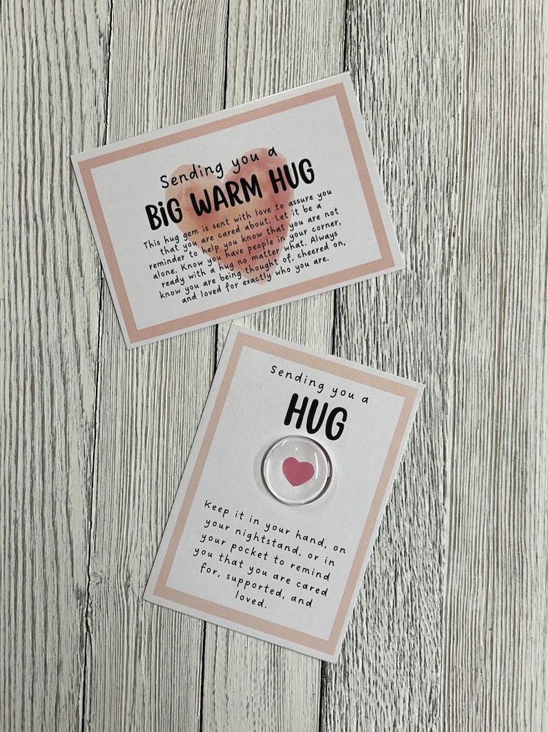 Puede incluir: Dos tarjetas con fondos de acuarela rosa. La tarjeta superior dice "Sending you a BIG WARM HUG" y la tarjeta inferior dice "Sending you a HUG". La tarjeta inferior tiene un peque&ntilde;o amuleto de coraz&oacute;n de vidrio con un coraz&oacute;n rosa en su interior.