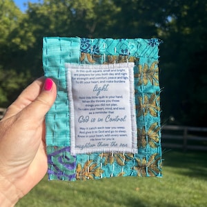 May include: A small, turquoise blue quilted square with a white label sewn on. The label has a poem printed on it that reads: "In this quilt square, small and bright, are prayers for you, both day and night For strength and comfort, peace and light, To lift your heart, and make burdens light Hold this little quilt in your hand, When life throws you those things you did not plan. To calm your heart, mind, and soul as a reminder that God is in Control May it catch each tear you weep. And give it to God and go to sleep. Know in your heart, with every seam His love for you is mightier than the sea"