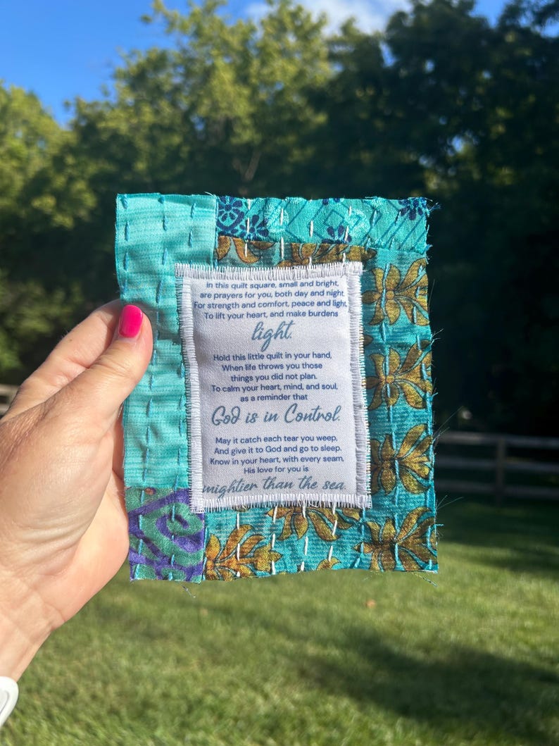 May include: A small, turquoise blue quilted square with a white label sewn on. The label has a poem printed on it that reads: "In this quilt square, small and bright, are prayers for you, both day and night For strength and comfort, peace and light, To lift your heart, and make burdens light Hold this little quilt in your hand, When life throws you those things you did not plan. To calm your heart, mind, and soul as a reminder that God is in Control May it catch each tear you weep. And give it to God and go to sleep. Know in your heart, with every seam His love for you is mightier than the sea"