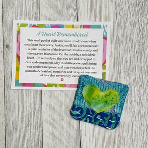 May include: A small blue and green fabric heart-shaped pocket quilt with a wooden heart inside. The quilt is accompanied by a card that reads "A Heart Remembered" and includes a message about the quilt's purpose as a reminder of love and comfort.