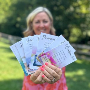 May include: A selection of inspirational cards with watercolour backgrounds and biblical text. The cards feature phrases from the Bible, including "2 Corinthians 5:17" and "Philippians." The cards are held in a person's hands.