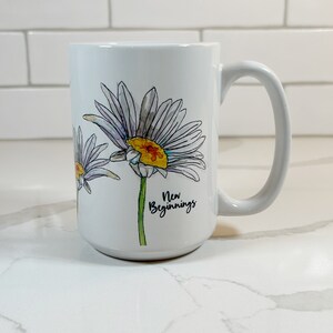 Pu&ograve; includere: Tazza in ceramica bianca con un'illustrazione ad acquerello di margherite e il testo "Nuovi Inizi".