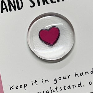 May include: A white card with the text "HOPE, COURAGE AND STRENGTH."  A small clear glass circle with a pink heart inside is attached to the card. The text "Keep it in your hand, your nightstand, or your pocket to remind you that you are strong." is written below the glass circle.