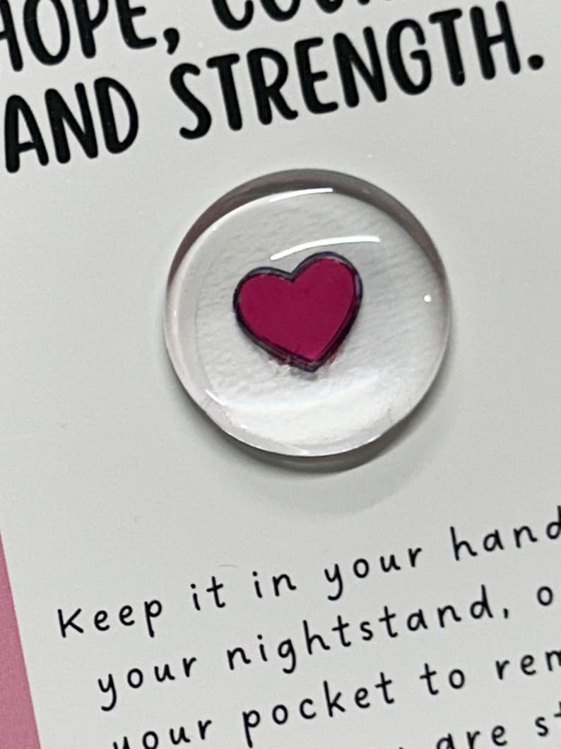 May include: A white card with the text "HOPE, COURAGE AND STRENGTH."  A small clear glass circle with a pink heart inside is attached to the card. The text "Keep it in your hand, your nightstand, or your pocket to remind you that you are strong." is written below the glass circle.
