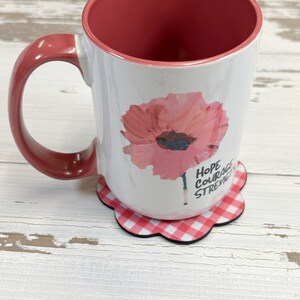 May include: White ceramic mug with a pink poppy flower design and the words "Hope Courage Strength" printed on the side. The mug is sitting on a red and white gingham coaster.