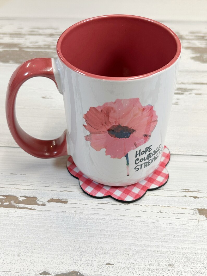 May include: White ceramic mug with a pink poppy flower design and the words "Hope Courage Strength" printed on the side. The mug is sitting on a red and white gingham coaster.