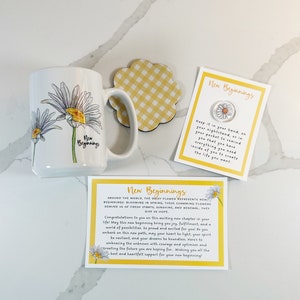 Pu&ograve; includere: Una tazza bianca con un manico giallo presenta un design con un fiore di margherita. La tazza &egrave; accompagnata da un sottobicchiere a quadri gialli e da un biglietto con il testo "Nuovi Inizi" e una grafica di margherita. Il biglietto recita "Tienilo in mano, sul tuo comodino o in tasca per ricordarti che hai tutto ci&ograve; di cui hai bisogno dentro di te per creare la vita che vuoi."