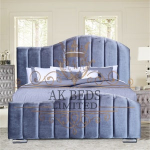 May include: A grey upholstered bed frame with a tall, curved headboard featuring vertical channel tufting. The headboard has a subtle wave pattern. The bed frame is accented with silver metal legs. The text "AK Beds Limited" is visible on the bed frame.