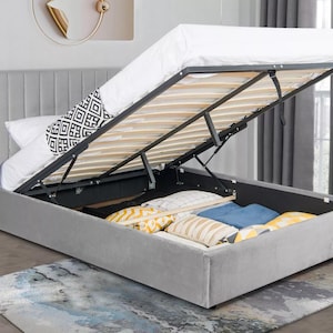 May include: A grey upholstered bed frame with a lift-up storage compartment. The bed frame has a tufted headboard and a slatted base. The storage compartment is open and reveals a black metal frame and a wooden slatted base.
