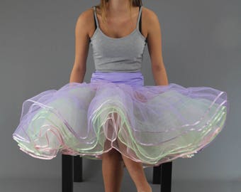 Puede incluir: Una persona joven que lleva un top gris sin mangas y una falda de tul de varias capas con detalles en rosa, verde y morado. La falda es muy amplia y tiene un aspecto de volantes. La persona está sentada en un taburete negro con los pies en el suelo.