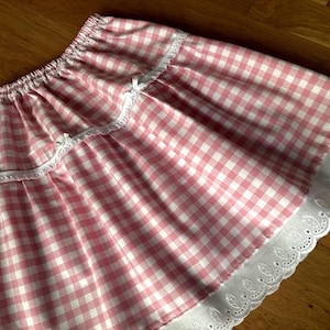 May include: A pink and white gingham skirt with a ruffled hem and white lace trim. The skirt has an elastic waistband and small white bows. The fabric is a lightweight cotton, perfect for warm weather.