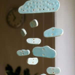 Può includere: Un mobile in ceramica blu chiaro con forme di nuvole appese a una corda. Le nuvole sono di diverse dimensioni e hanno piccoli fori.