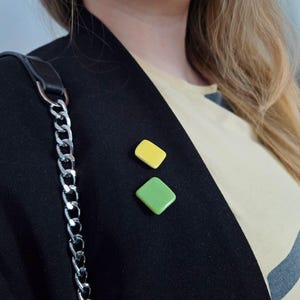 Op de afbeelding: Een zwarte blazer versierd met twee vierkante spelden: een gele en een groene. Een zilveren kettingriem hangt aan de linkerkant. De blazer wordt gedragen over een lichtgekleurd shirt met een donker geometrisch ontwerp.