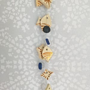 May include: A cream-coloured, handmade mobile with geometric shapes and starburst designs. The mobile features various shapes, including diamond, leaf, and star forms, strung together with a clear thread. Accents include gold and blue details.