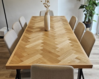 May include: A wooden dining table with a herringbone pattern top and black metal legs. Six chairs with beige fabric upholstery surround the table. A vase with dried flowers sits in the center of the table.