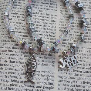 May include: A necklace featuring clear and iridescent beads, with silver-toned geometric beads and a pendant with the word "JESUS" in a fish shape. The necklace is displayed on an open book.
