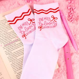 May include: White socks with red detailing and the pink text "ENEMIES to lovers BOOKCLUB". The socks feature a wavy red pattern at the top and a bow graphic. A pink pen and book are in the background.