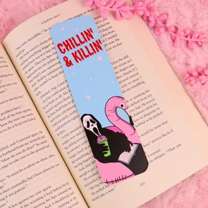 Op de afbeelding: Een rechthoekige boekenlegger met een lichtblauwe achtergrond, kleine roze sterretjes en de rode tekst CHILLIN' & KILLIN'. Het ontwerp toont een in zwart gehulde figuur met een wit masker die een groen drankje en een boek vasthoudt, naast een roze flamingo-zwemband.