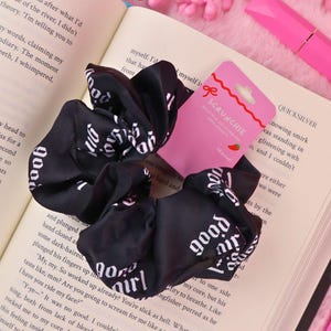 Könnte beinhalten: Schwarzes Haargummi mit weißem "good girl" Schriftzug. Hergestellt aus seidigem Material, liegt es auf einem offenen Buch. Ein rosa Etikett mit dem Wort "SCRUNCHIE" ist sichtbar.