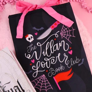 May include: Black book sleeve with a pink bow, featuring the text "The Villain Lover Book Club." The design includes a skull, hearts, bats, a knife, and spiderwebs. The book sleeve is designed to protect books.