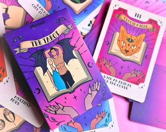 May include: A purple and pink deck of cards called "TBR Tarot" with a couple on the cover. The couple is standing in front of an open book with a bat's wings behind them. The cards are on a pink surface.