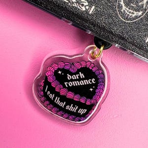 Peut inclure: Un porte-clés en forme de cœur transparent avec un cœur noir à l'intérieur. Le cœur porte l'inscription "dark romance" et "I eat that shit up". Le porte-clés est rose et violet.