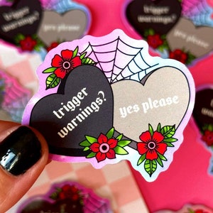 Peut inclure: Deux autocollants en forme de cœur avec un motif de toile d'araignée. Un cœur est noir avec le texte "trigger warnings?" et l'autre est gris avec le texte "oui s'il vous plaît".