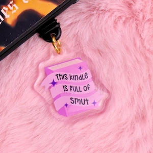 Könnte beinhalten: Ein klarer Acryl-Anhänger mit einem goldfarbenen Ring. Der Anhänger hat die Form eines Buches mit dem Text "This kindle IS FULL OF SMUT" in Lila. Der Anhänger hängt vor einem rosa, flauschigen Hintergrund.