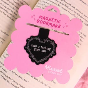 Op de afbeelding: Roze magnetische bladwijzer met een geschulpte rand. De bladwijzer heeft de tekst "MAGNETIC BOOKMARK" en "such a fucking good girl" in een hartvorm. De bladwijzer ligt op een open boek.