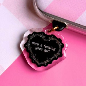 Op de afbeelding: Een doorzichtige telefoonbedel met een zwart hartvormig ontwerp met prikkeldraad en de tekst "such a fucking good girl" in het wit. De bedel is bevestigd aan een telefoonhoesje met een goudkleurige ring.