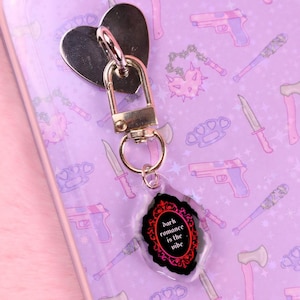 May include: A silver heart-shaped keychain with a gold clasp and a clear charm. The charm has the text "dark romance is the vibe" in a black and red ornate frame. The background is a light purple with weapon illustrations.