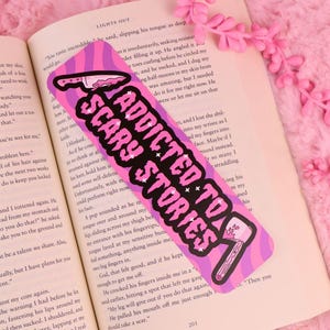 May include: A rectangular bookmark with a pink and purple wavy background, featuring the text "ADDICTED TO SCARY STORIES" in a bold, white, bubbly font, accented by a graphic of a knife at the top and a bloody axe at the bottom.
