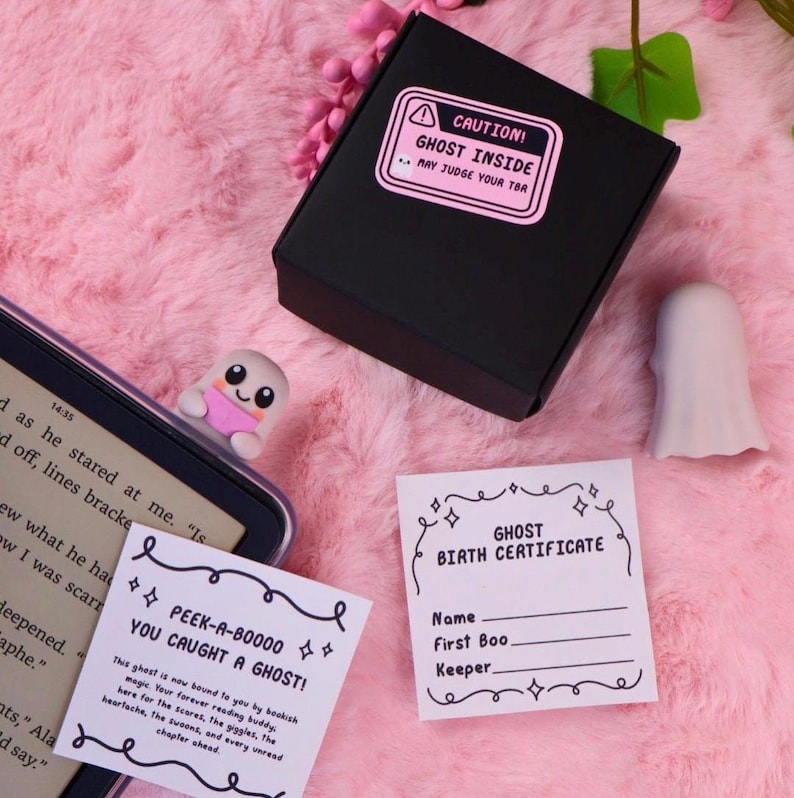May include: A black box with a pink label that reads "CAUTION! GHOST INSIDE." A ghost-shaped bookmark, a ghost-shaped figurine, and two cards with ghost-themed text are displayed on a pink, fluffy surface.