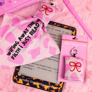Op de afbeelding: Roze leesaccessoires, waaronder een schermreinigingsdoekje met een rode strik, een roze en wit patroon doek met tekst en een roze stylus. Een e-reader met een schildpadframe is ook zichtbaar.