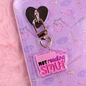 May include: A heart-shaped metal keychain with a lobster clasp and a clear acrylic tag. The tag reads "DEFINITELY NOT reading SMUT" in pink and black lettering. The background is a purple case with weapon illustrations.