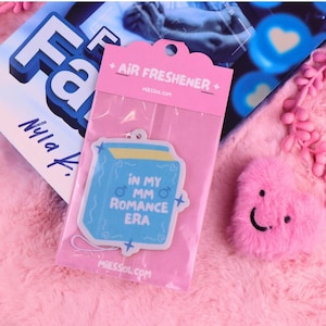 May include: A pink air freshener in a clear plastic package with the text "AIR FRESHENER" and "MIESSOL.COM". The air freshener is light blue with the text "IN MY OMM ROMANCE ERA". A pink fluffy heart is also visible.