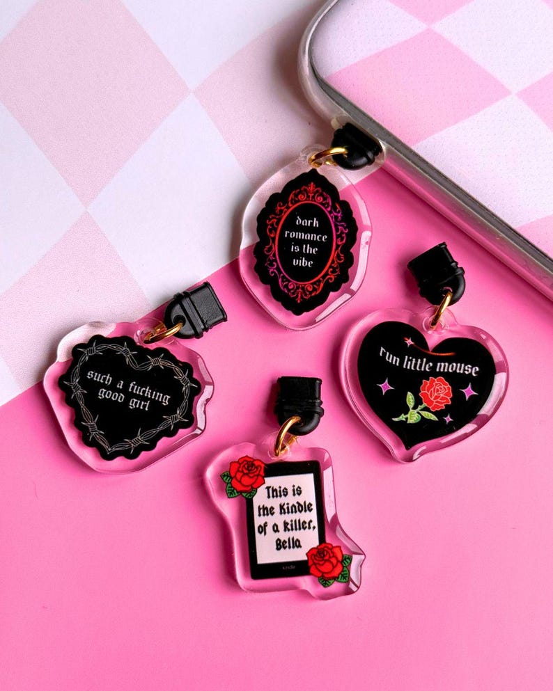K&ouml;nnte beinhalten: Vier Acryl-Anh&auml;nger mit schwarzem Text und goldfarbener Hardware. Die Anh&auml;nger haben die Form eines Herzens, einer Flasche und einer dekorativen Form. Der Text lautet "dark romance is the vibe", "such a fucking good girl", "Fun little mouse" und "This is the Kindle of a killer, Bella".
