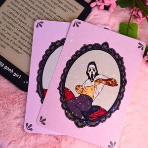 Op de afbeelding: Twee roze speelkaarten met een glitterafwerking. Elke kaart heeft een omlijste illustratie van een personage uit de Scream-filmreeks. De kaarten zijn versierd met zwart filigraan en kleine bloemaccenten.