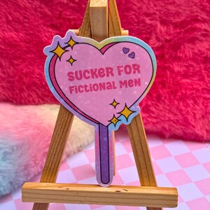 May include: A pink heart-shaped lollipop sticker with a rainbow border and glitter. The sticker says "SUCKER FOR FICTIONAL MEN" in white text. The lollipop is on a small wooden easel.