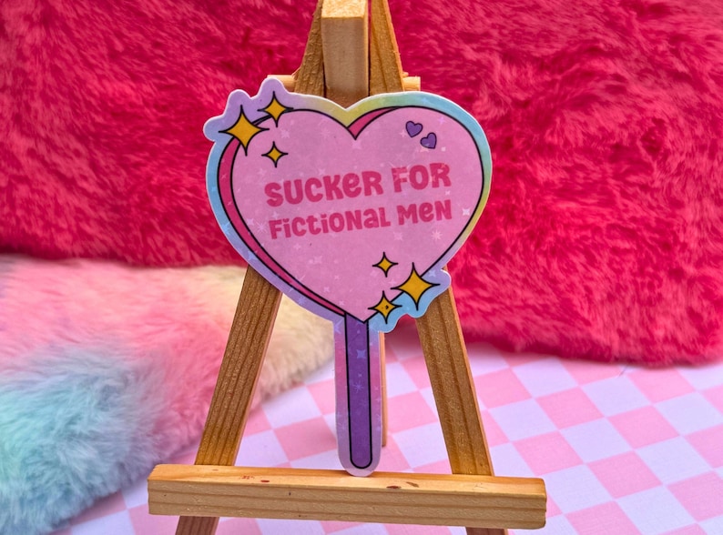 May include: A pink heart-shaped lollipop sticker with a rainbow border and glitter. The sticker says "SUCKER FOR FICTIONAL MEN" in white text. The lollipop is on a small wooden easel.