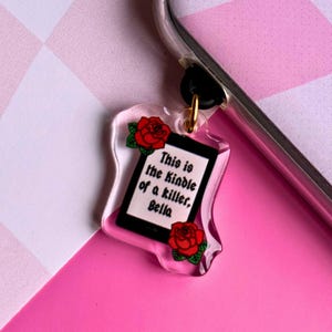 May include: A clear plastic charm with a black and white image of a Kindle and the text "This is the Kindle of a killer, Bella". The charm is decorated with red roses.