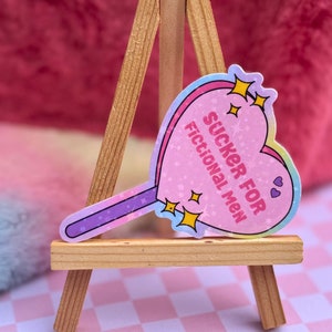 May include: A pink heart-shaped lollipop sticker with the text "SUCKER FOR FICTIONAL MEN" and glitter accents. The sticker is on a wooden easel.