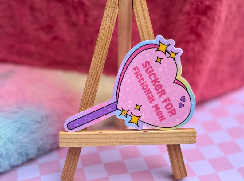 May include: A pink heart-shaped lollipop sticker with the text "SUCKER FOR FICTIONAL MEN" and glitter accents. The sticker is on a wooden easel.