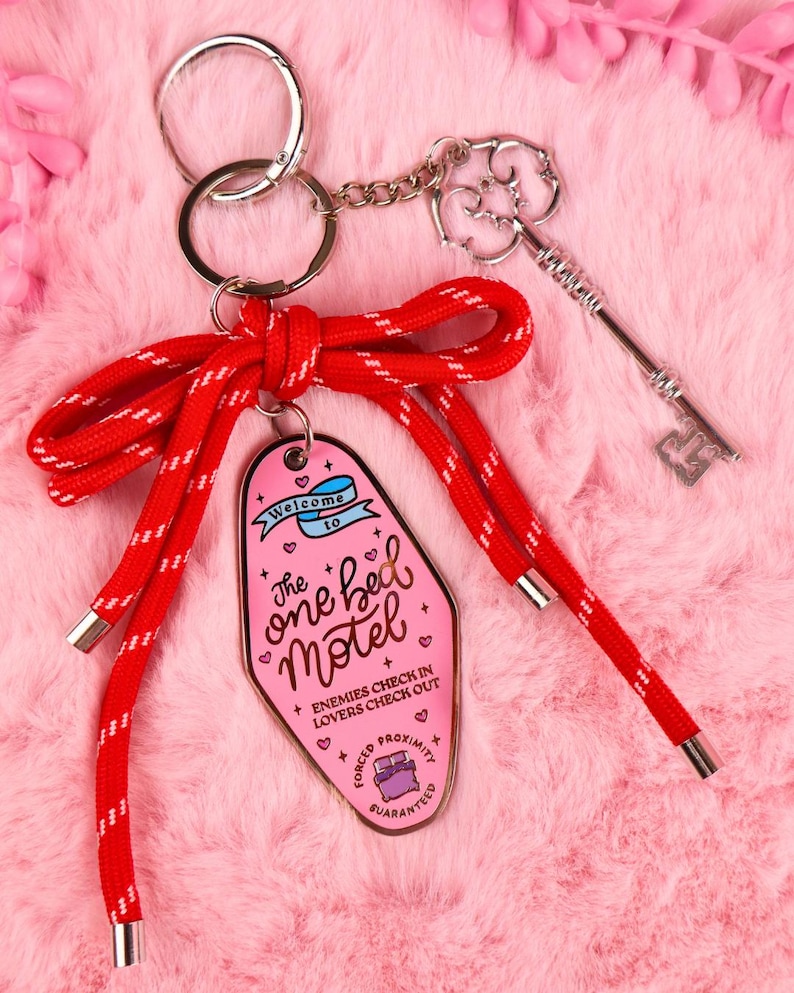 Puede incluir: Un llavero rosa estilo motel con el texto "The one bed Motel" y un cord&oacute;n rojo. El llavero incluye una llave plateada y una cadena. El texto en el llavero dice "Enemies check in, Lovers check out, Forced Proximity Guaranteed."
