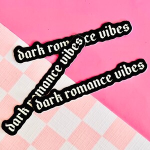 May include: Three black stickers with white text that reads "dark romance vibes" on a pink and white checkered background.
