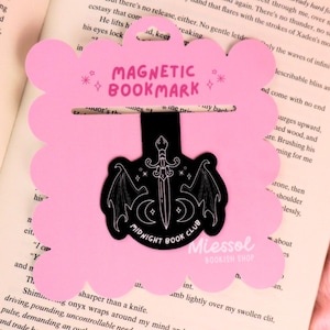 May include: A pink magnetic bookmark with a scalloped edge. The bookmark has the text "MAGNETIC BOOKMARK" and a black design of a sword, wings, and moons. The text "MIDNIGHT BOOK CLUB" is also visible.