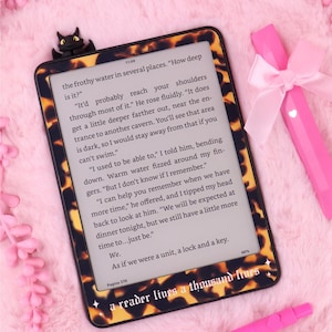 May include: An e-reader with a tortoiseshell pattern case, displaying text from a book. A black cat-shaped bookmark sits at the top. A pink stylus and a pink bow are on the side. The text on the e-reader says "a reader lives a thousand lives".