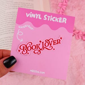 May include: A pink vinyl sticker with the words "BOOK LOVER" in red script. The sticker is on a pink card with the words "VINYL STICKER" at the top and "WATERPROOF!" below. The website address MIESSOL.COM is at the bottom.