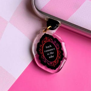 May include: A clear, oval-shaped phone charm with a black background and a gold-colored metal ring. The charm features a decorative black frame with a red and pink gradient and the text "dark romance is the vibe" in white.
