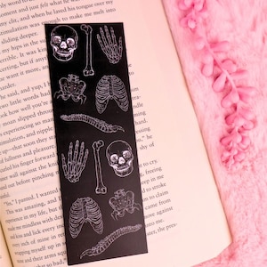 Op de afbeelding: Zwarte boekenlegger met witte anatomische illustraties van botten, waaronder een schedel, ribbenkast en wervelkolom. De boekenlegger is rechthoekig en ligt op een open boek.